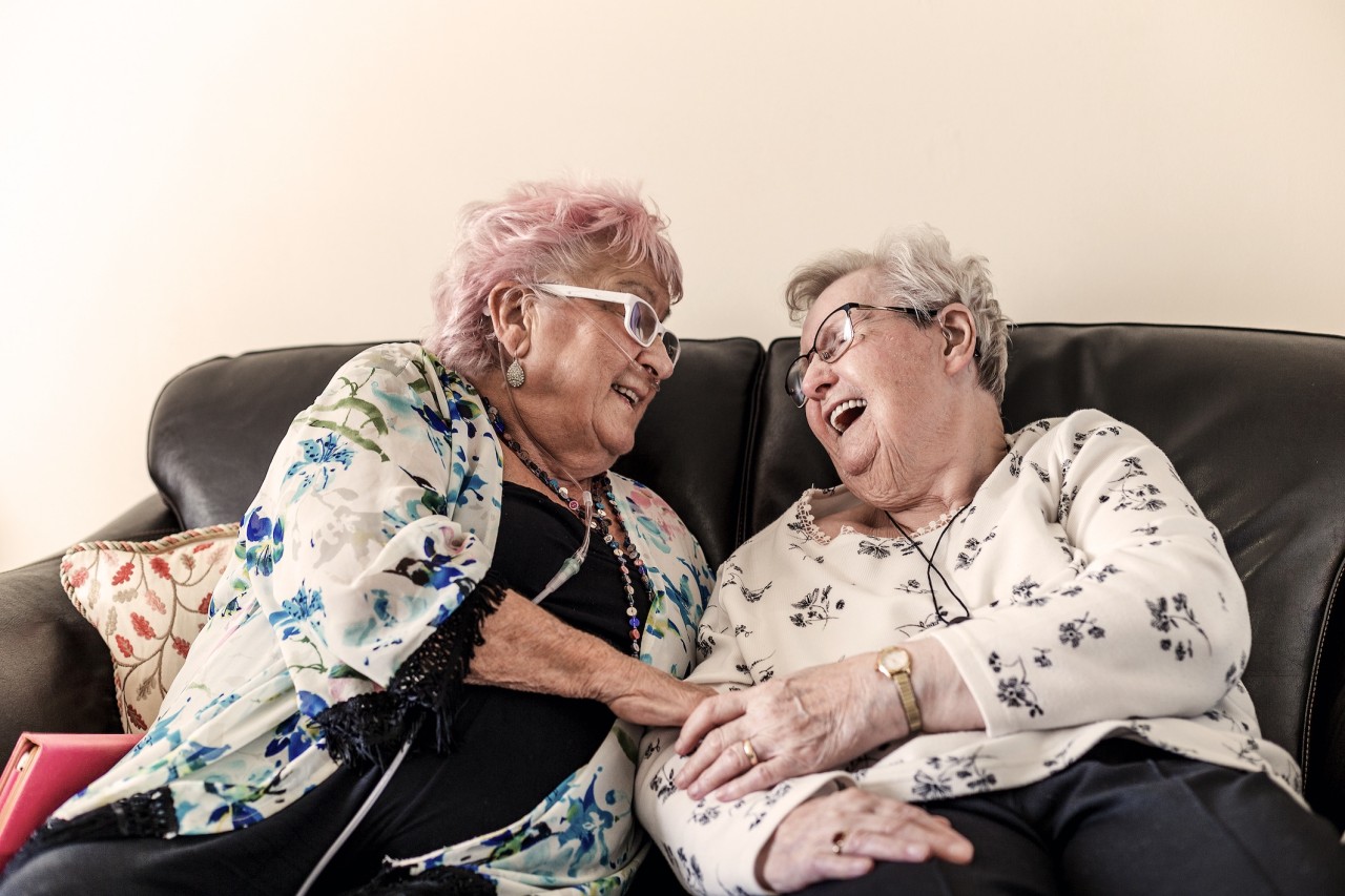 Twee oudere dames zitten lachend op de bank en houden elkaars hand vast